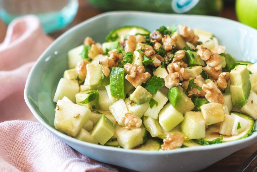 A modern light blue bowl containing a delicious apple salad with raw courgette, avocado, walnuts, EVOO, balsamic vinegar, salt and pepper. In the background, courgettes and a glass of lemon water can be seen. Everything is fitness.