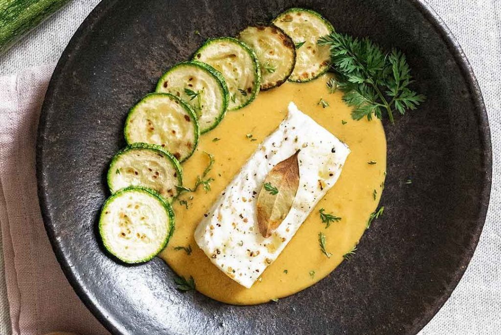 Plato de piedra sobre un trapo de cocina rosa, presentando bacalao al horno con una salsa cocinada de color naranja y una hoja de decoración encima, rodeado por siete rodajas de calabacín a la plancha que añaden un toque colorido y apetitoso al plato, decorado finalmente con un poco de perejil.