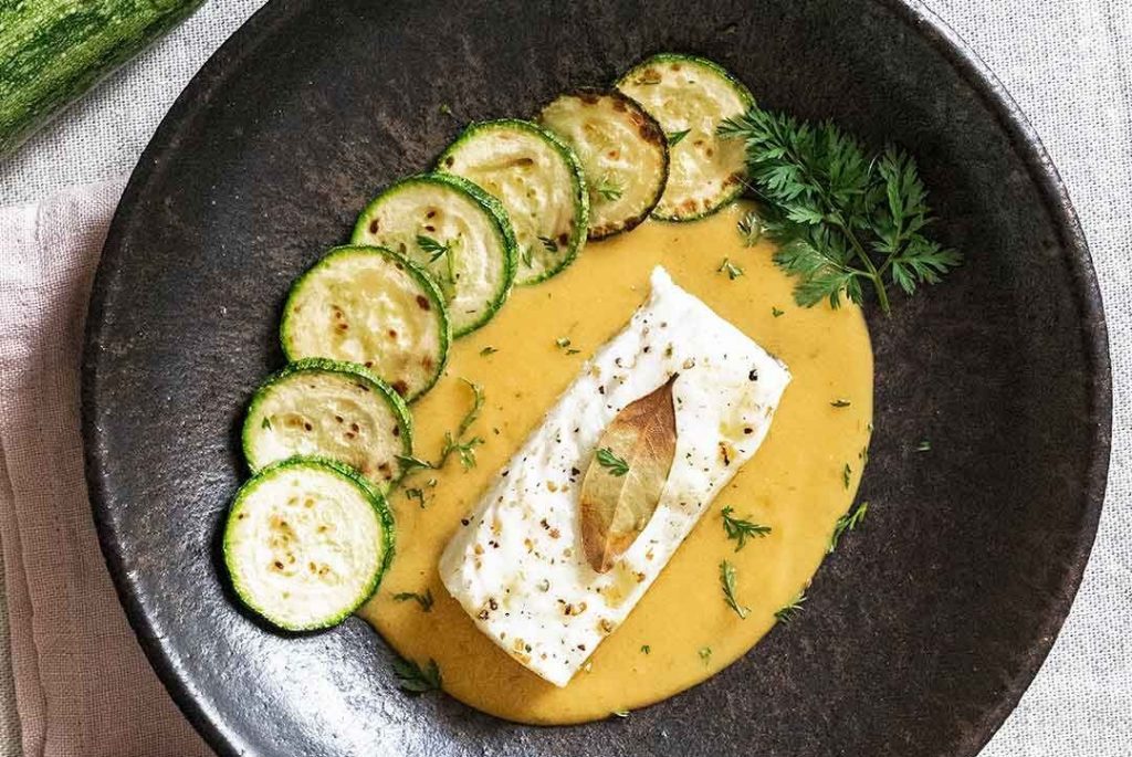 Plat de pedra sobre un drap de cuina rosa, presentant bacallà al forn amb una salsa cuinada de color taronja i una fulla de decoració a sobre, envoltat per set rodanxes de carbassa a la planxa que afegeixen un toc colorit i apetitós al plat, finalment decorat amb una mica de julivert.