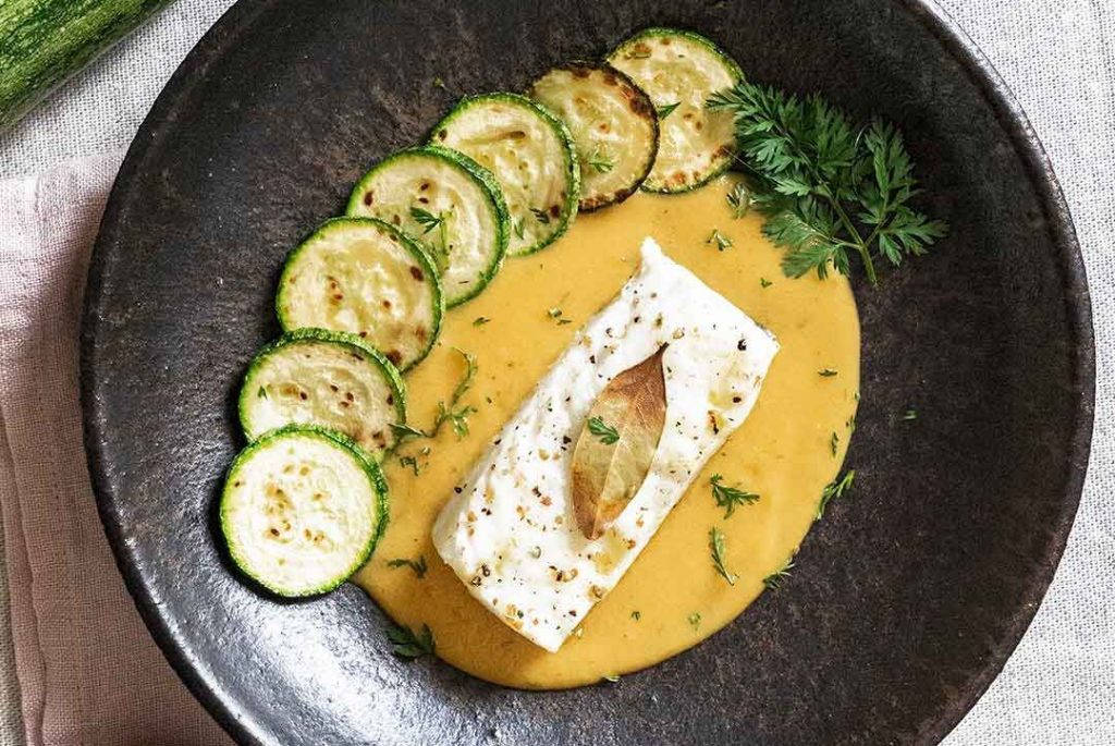 Stone plate on a pink kitchen towel, presenting baked cod with a cooked orange sauce and a decoration leaf on top, surrounded by seven grilled courgette slices that add a colorful and appetizing touch to the dish, finally decorated with a bit of parsley.