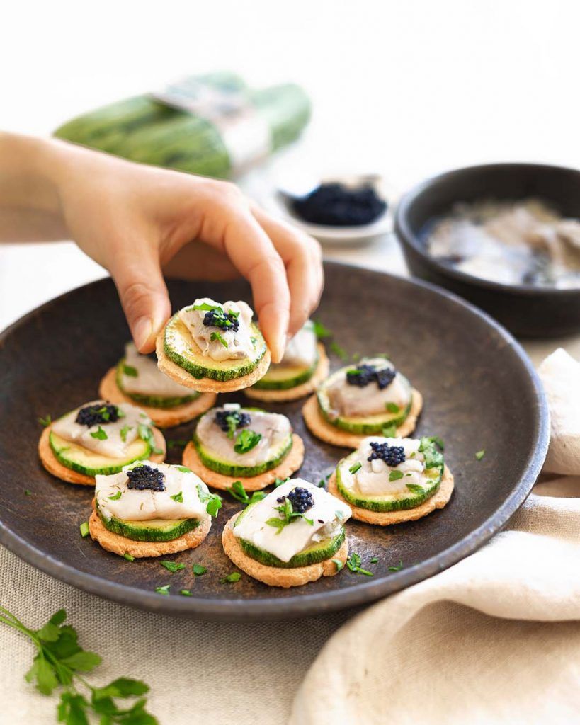 Plato hondo de piedra lleno de canapés elaborados con calabacín crudo, arenque marinado, huevas de pescado y perejil. Una mano presenta uno de los canapés. Al fondo, se ven calabacines y los ingredientes para la elaboración del plato difuminados. Estos canapés son una forma perfecta de comerse la vida de un bocado.