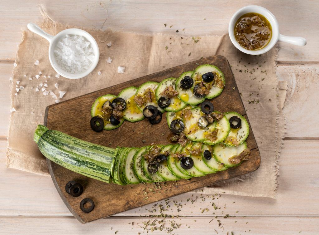 Una taula de cuina sobre un drap antic presenta carbassa tallada a rodanxes, adornada amb olives negres, sal, oli d'oliva verge extra, orenga i salsa de mostassa. Un plat fresc i saludable de carpaccio de carbassa que combina textures i sabors per a una experiència culinària deliciosa.