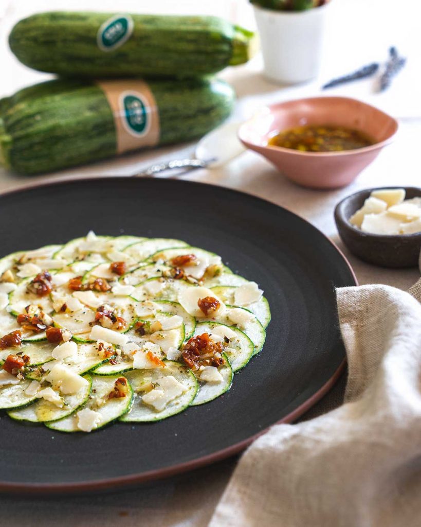 Assiette ronde sombre avec du carpaccio de courgette crue coupée en rondelles, avec du fromage parmesan, de l'huile d'olive, des tomates séchées, des noisettes, du sel et du poivre. Une délicieuse solution à l'absence de temps pour faire les courses.