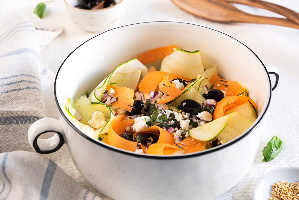 Un bol blanco con asas que parece una olla de porcelana, contiene calabacín crudo, zanahoria, tomates cherry, aceitunas negras y queso feta, listo para mezclar con tu salsa favorita.