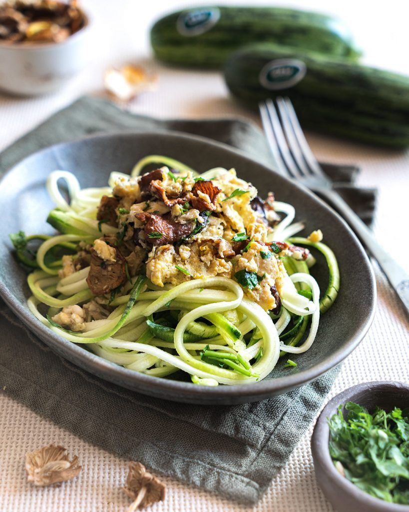 Un plato hondo gris lleno de espaguetis de calabacín coronados con setas y huevo, con un aspecto muy apetitoso. Alrededor del plato, hay cuencos con la salsa que los acompaña y unos calabacines. La pasta de calabacín se realza con el sabor de las setas y el huevo.