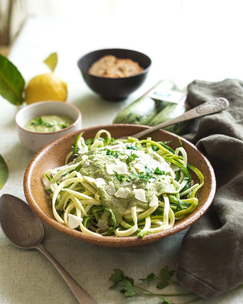 Un bol de color barro lleno de espaguetis con salsa de brócoli y calabacín crudo, espolvoreado con parmesano y con un aspecto delicioso. Alrededor del plato, hay cuencos con la salsa que los acompaña y algunas tostadas. La pasta se enriquece con el sabor fresco del calabacín crudo y la salsa de brócoli.