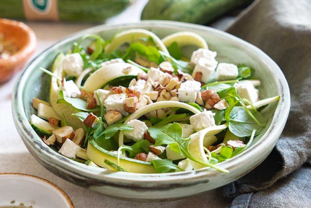 A round bowl on a kitchen towel containing raw courgette, feta cheese, hazelnuts, rocket, EVOO, salt, pepper, vinegar, chopped coriander, and lemon. In the background, blurred raw courgettes can be seen.