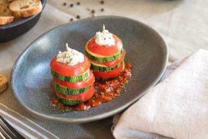 Two skewers with alternating slices of courgette and tomato, topped with burrata and seasoned with basil, presented on an oval imitation stone deep plate, on a tomato sauce.