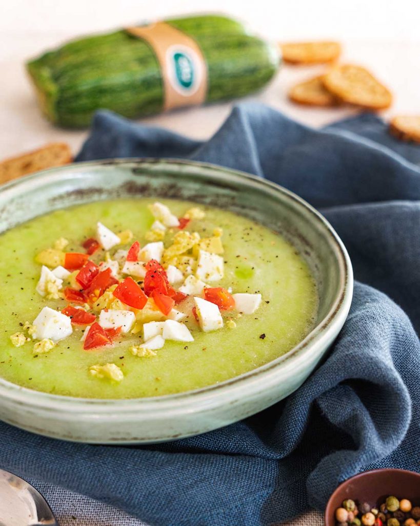 Bol rond blanc rempli de gaspacho aux courgettes crues, décoré de morceaux d'oeuf et de tomate, donnant au plat une couleur exceptionnelle et une apparence appétissante, présenté sur un torchon de cuisine sur un comptoir, avec deux courgettes floues en arrière-plan comme ingrédient principal de ce plat.
