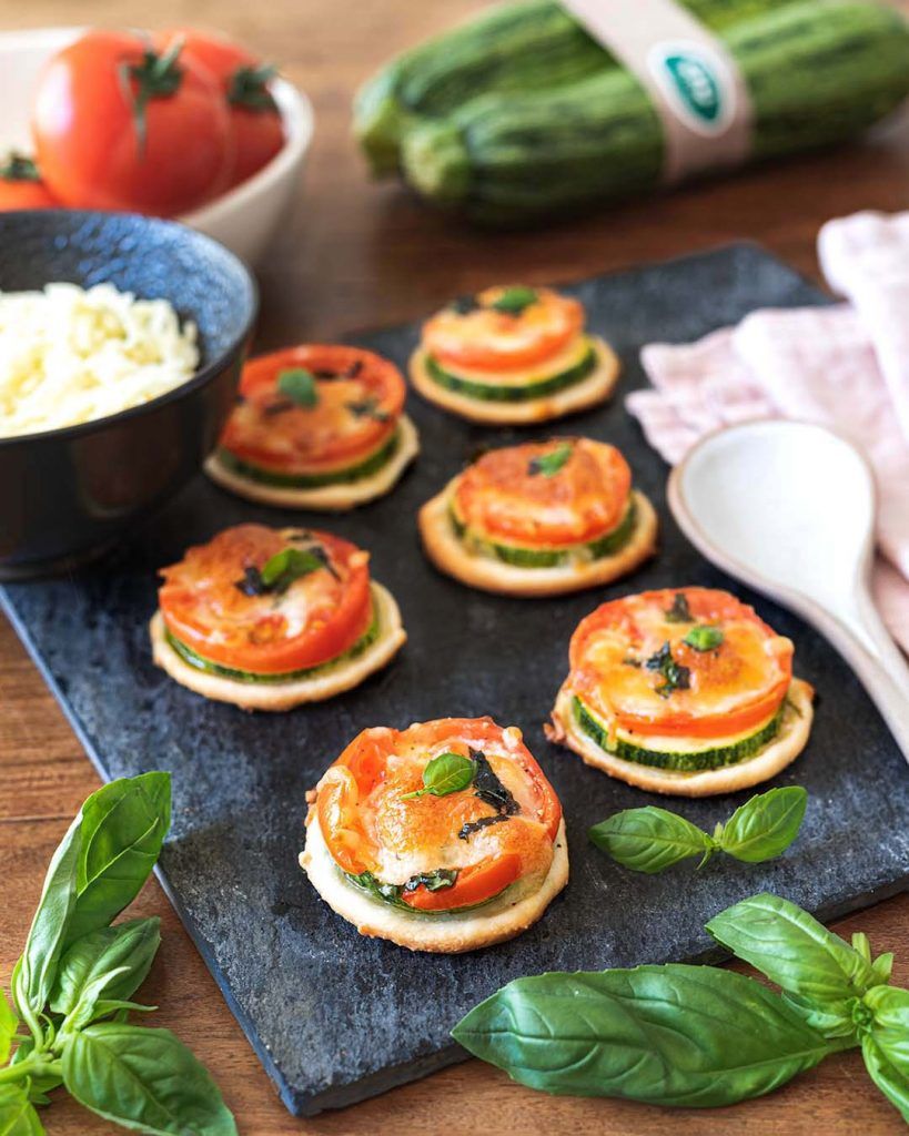 Plusieurs mini pizzas à la courgette crue disposées sur une belle plaque d'ardoise noire. On voit plusieurs bols avec les ingrédients autour, une grande cuillère et un joli torchon de cuisine. Ces mini pizzas sont une façon parfaite de savourer la belle Italie en une seule bouchée.