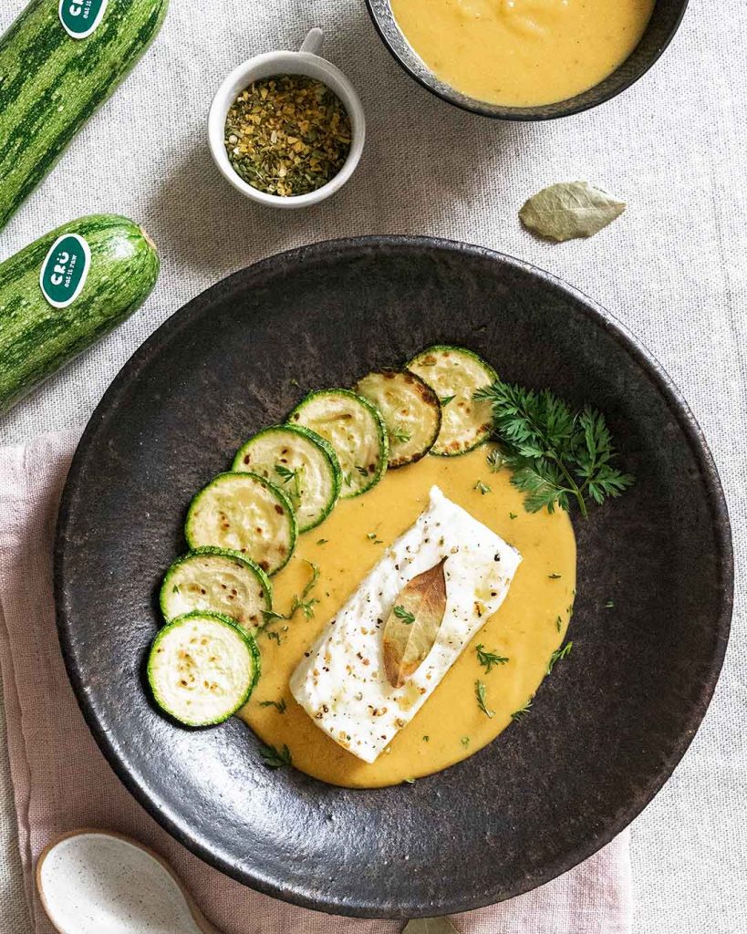 Assiette en pierre sur un torchon de cuisine rose, présentant de la morue au four avec une sauce cuisinée de couleur orange et une feuille de décoration sur le dessus, entourée de sept tranches de courgettes grillées qui ajoutent une touche colorée et appétissante au plat, finalement décorée avec un peu de persil.