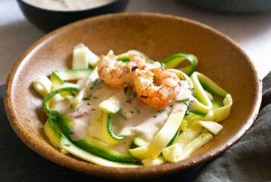 A bowl full of noodles with shrimp and raw courgette, looking delicious. Around the dish, there are bowls with the accompanying sauce and some toast. The noodles are a crunchy and healthy way to enjoy pasta, without giving up flavor.