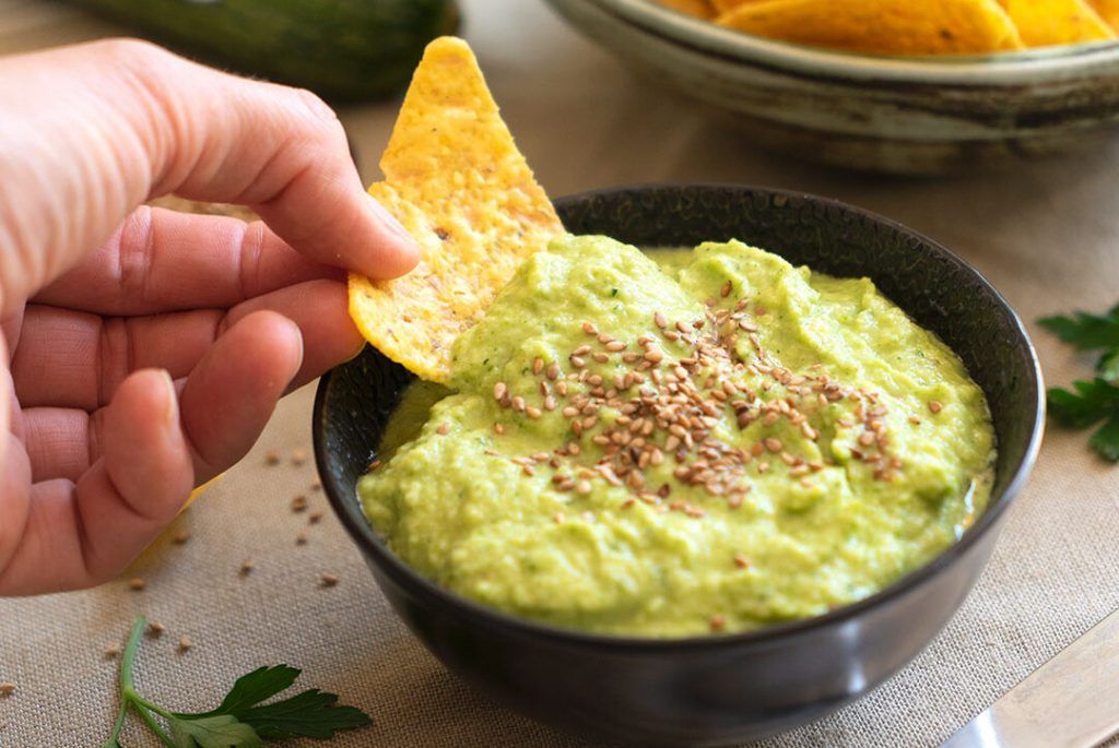 Un pâté de courgette cru qui ressemble à un guacamole, par sa couleur verte et parce qu'il est présenté dans un petit bol rond et sombre, avec un Dorito prenant un peu. Il a des graines sur le dessus pour la décoration. Vert que je t'aime vert et si c'est du guacamole de courgette crue, encore plus! Notez cette recette fitness!
