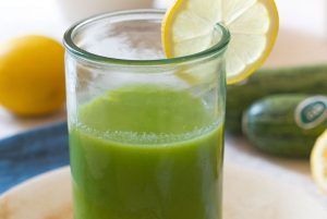 A transparent glass containing a striking, intense green smoothie made from raw courgette, apple and lemon, decorated with a slice of lemon. In the background, courgettes and lemons can be seen. This smoothie, served on a plate with a spoon, is the perfect recipe for all those who dislike green and will only take you 5 minutes to prepare.