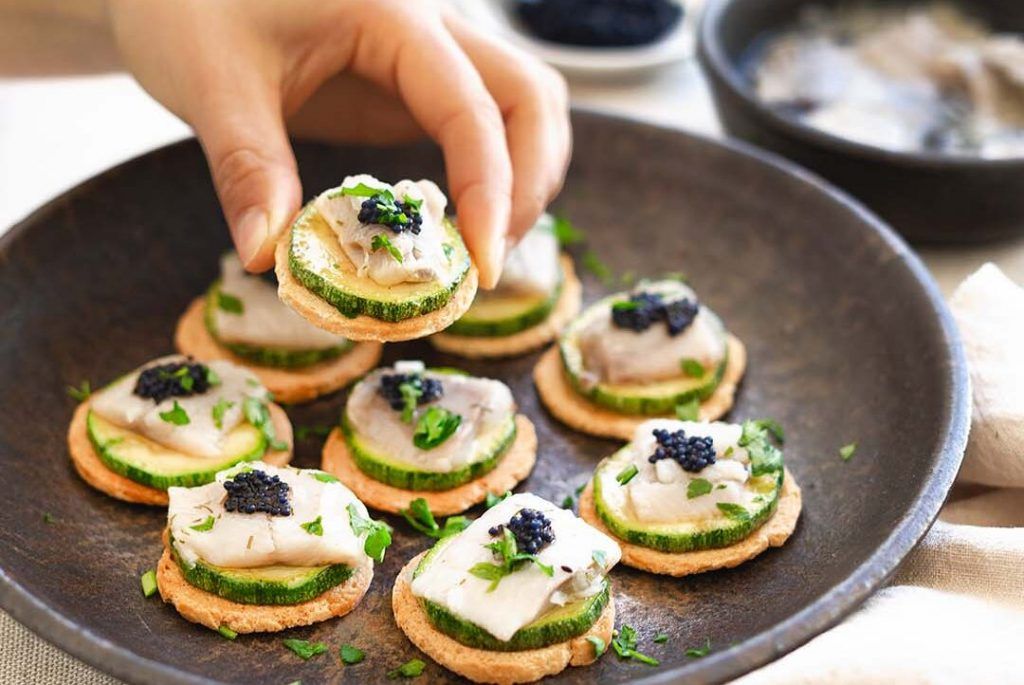 Stone deep dish full of canapés made with raw courgette, marinated herring, fish roe and parsley. A hand is presenting one of the canapés. In the background, courgettes and the ingredients for the dish preparation are seen blurred. These canapés are a perfect way to eat life in one bite.