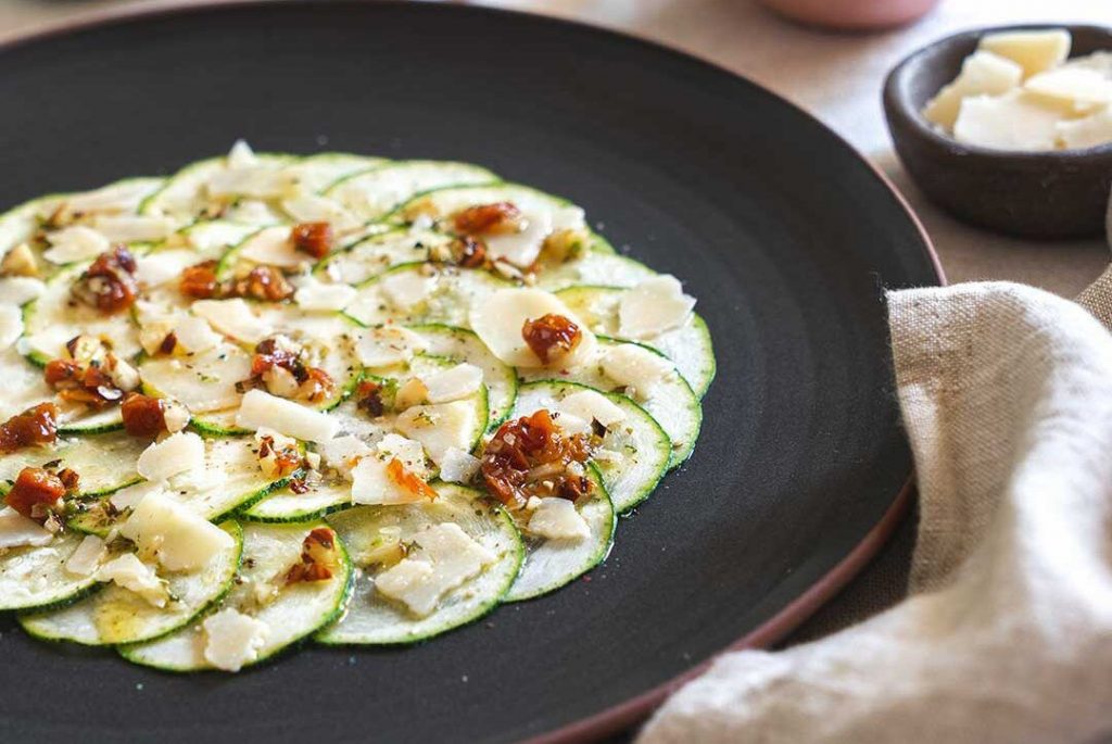 Dark round plate with raw courgette carpaccio cut into slices, with parmesan cheese, olive oil, dried tomato, hazelnuts, salt, and pepper. A delicious solution to the lack of time for shopping.