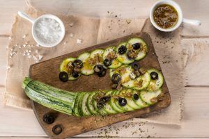 A kitchen board on an antique cloth presents courgette sliced, garnished with black olives, salt, extra virgin olive oil, oregano, and mustard sauce. A fresh and healthy courgette carpaccio dish that combines textures and flavors for a delicious culinary experience.