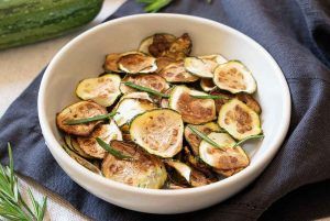 A bowl with slices of raw courgette fried as chips in the air fryer, presented on a kitchen towel and decorated with rosemary around. Try the modern and cool method of the air fryer to enjoy these courgette chips.