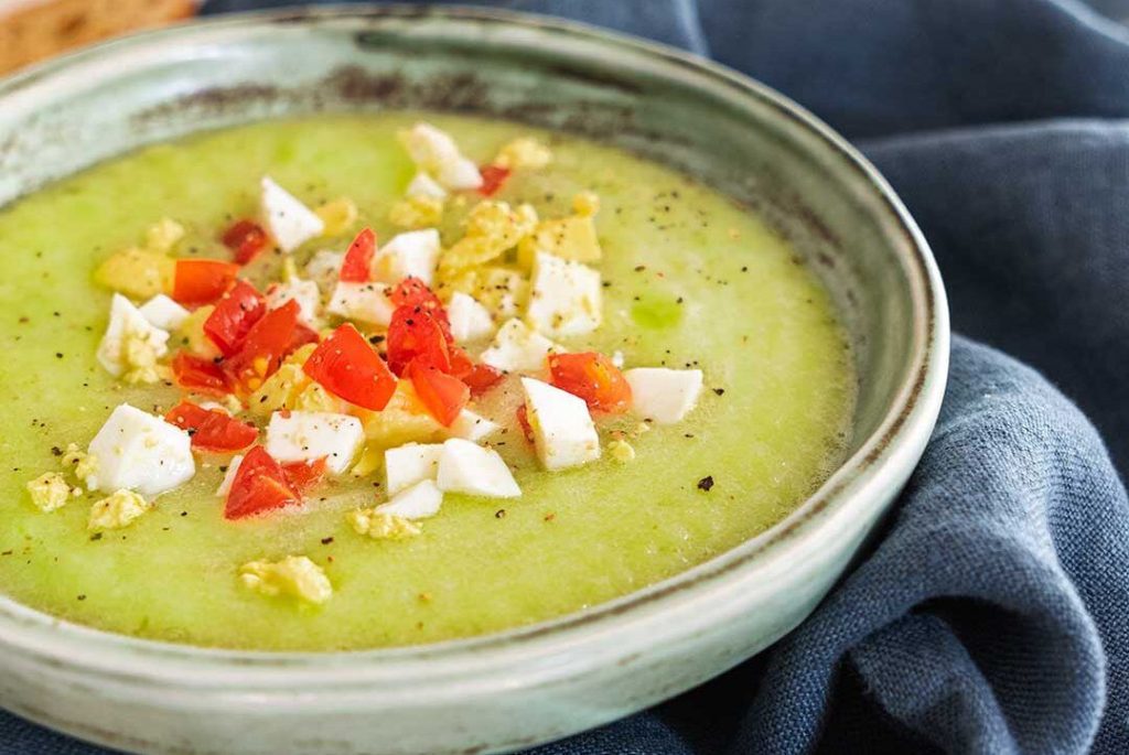 White round bowl filled with raw courgette gazpacho, garnished with bits of egg and tomato, giving the dish an exceptional color and an appetizing look, presented on a kitchen cloth on a countertop, with two blurred courgettes in the background as the main ingredient of this dish.