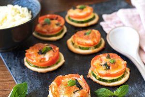Several one-bite mini pizzas with raw courgette arranged on an attractive black slate piece. There are several bowls with the ingredients around, a large spoon, and a very cute kitchen cloth. These mini pizzas are a perfect way to savor beautiful Italy in just one bite.