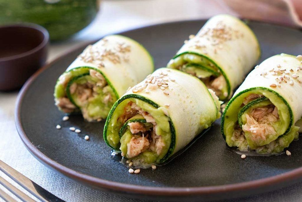 Round plate with four raw courgette rolls filled with guacamole and tuna. Decorated with olive oil, chopped parsley, chopped tomato and seeds. A delicious and fun bite without the drama of a soap opera.