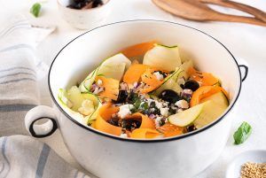 A white bowl with handles that looks like a porcelain pot, contains raw courgette, carrot, cherry tomatoes, black olives, and feta cheese, ready to mix with your favorite sauce.