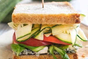 An appetizing sandwich with raw courgette, toasted bread, tomato, lamb's lettuce and cheese, presented on a flat plate. In the background, blurred courgettes can be seen.