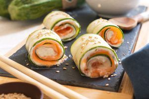 Four sushi rolls made with fresh courgette, spreadable cheese, salmon, seeds and soy sauce, presented on a slate board with Japanese chopsticks. A quick and delicious recipe that takes sushi to another level.