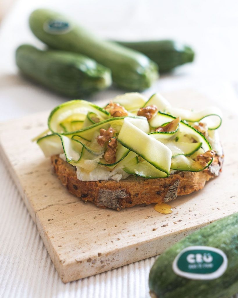 Imatge de la recepta "Bruschetta amb Nous", mostrant una torrada integral untada amb formatge i decorada amb rodanxes primes de carbassó. Les nous picades es col·loquen sobre el carbassó, afegint un toc de sabor i textura al plat.