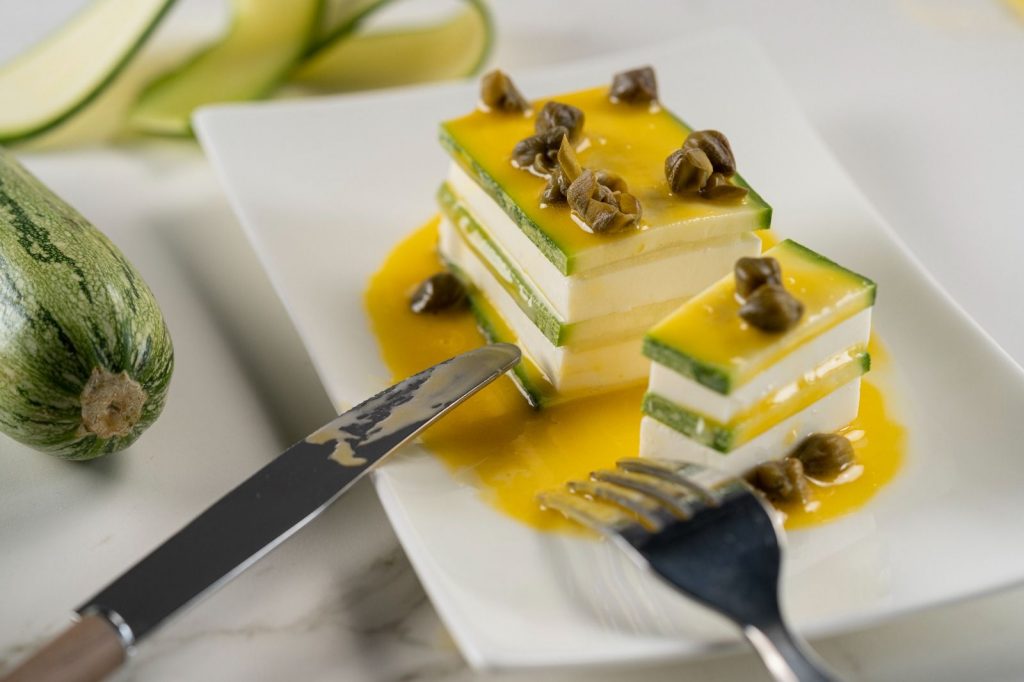 Imagen de la receta "Milhojas de Calabacín", que muestra una presentación elegante del plato con capas de calabacín y trozos de queso, bañados en aceite de trufa. En la parte superior se encuentran tres alcaparras como decoración. El plato está colocado sobre una pieza blanca y se muestra un tenedor y un cuchillo, como si acabaran de probar el delicioso plato.