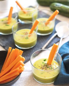 Image of the "Cream Shots" recipe, showing small elegant glasses filled with a soft cream made of Crü zucchini, leek, potato, carrots, vegetable broth, cooking cream of oats, extra virgin olive oil, salt, pepper and nutmeg. Each glass is garnished with a slice of carrot as a snack. This recipe, presented in an attractive and appetizing way, is perfect for those who miss their grandmother's homemade cream.