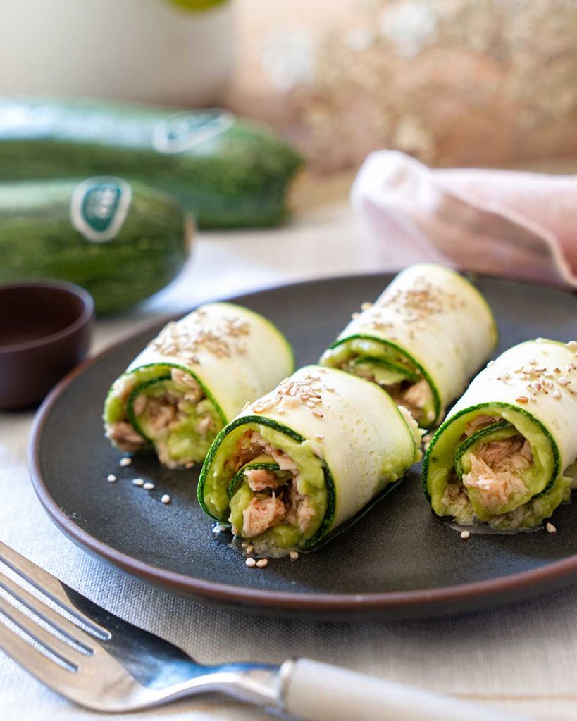 Assiette ronde avec quatre rouleaux de courgette crue farcis de guacamole et de thon. Décorés avec de l'huile d'olive, du persil haché, des tomates hachées et des graines. Une délicieuse bouchée amusante sans le drame d'un feuilleton.