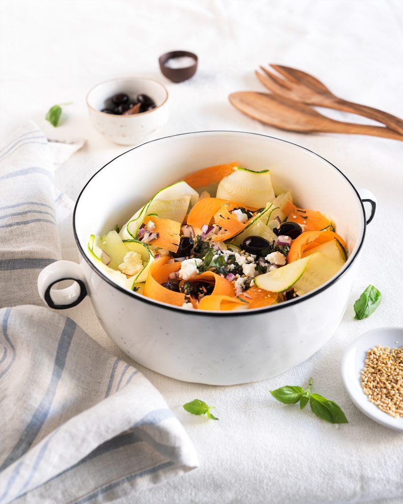 Un bol blanc avec des poignées qui ressemble à une marmite en porcelaine, contient des courgettes crues, des carottes, des tomates cerises, des olives noires et du fromage feta, prêt à être mélangé avec votre sauce préférée.