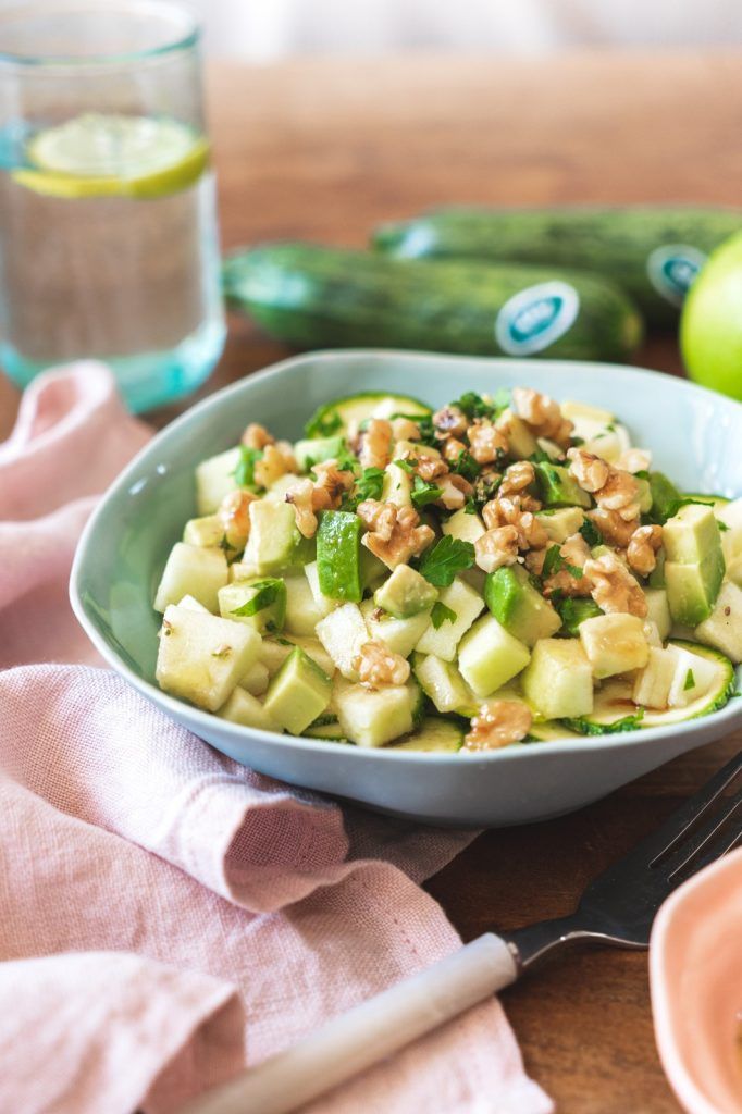 Un bol bleu clair moderne contenant une délicieuse salade de pommes avec des courgettes crues, de l'avocat, des noix, de l'huile d'olive, du vinaigre balsamique, du sel et du poivre. En arrière-plan, on voit des courgettes et un verre d'eau citronnée. Tout est fitness.