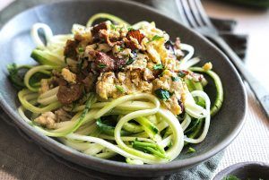 A grey deep plate full of courgette spaghetti crowned with mushrooms and egg, looking very appetising. Around the dish, there are bowls with the accompanying sauce and some courgettes. The courgette pasta is enhanced with the taste of mushrooms and egg.