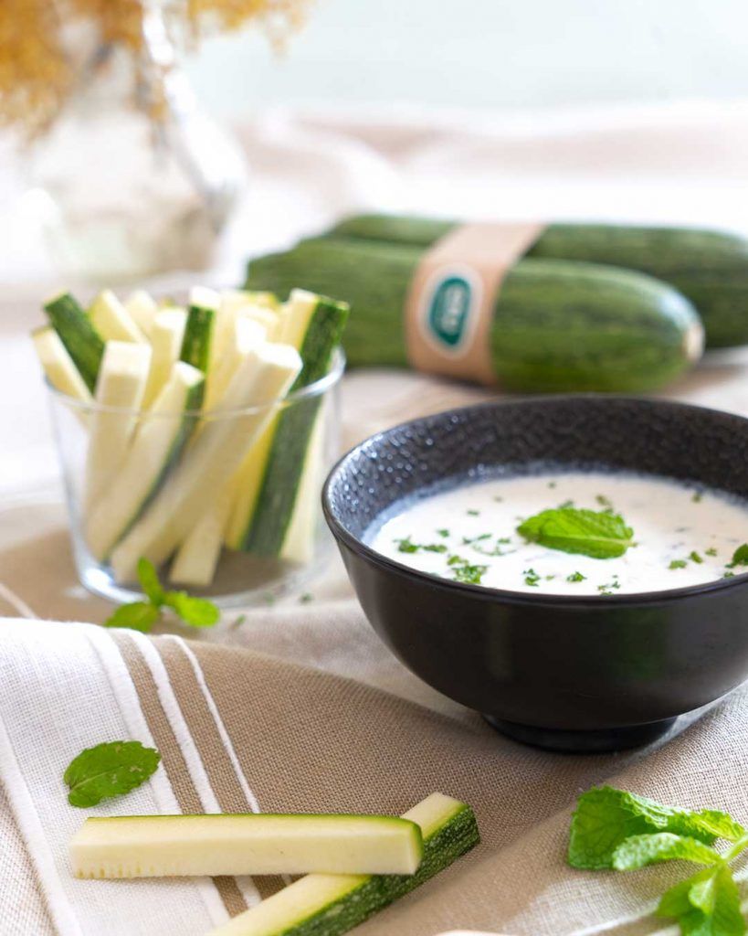 Un bol amb salsa tzatziki i un got amb tires de carbassa fresca com a snack. Una recepta fà cil i deliciosa que combina la cremositat de la salsa tzatziki casolana amb la textura cruixent de les tires de carbassa.