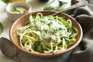 A clay-colored bowl full of spaghetti with broccoli sauce and raw courgette, sprinkled with Parmesan and looking delicious. Around the dish, there are bowls with the accompanying sauce and some toast. The pasta is enhanced with the fresh taste of raw courgette and broccoli sauce.