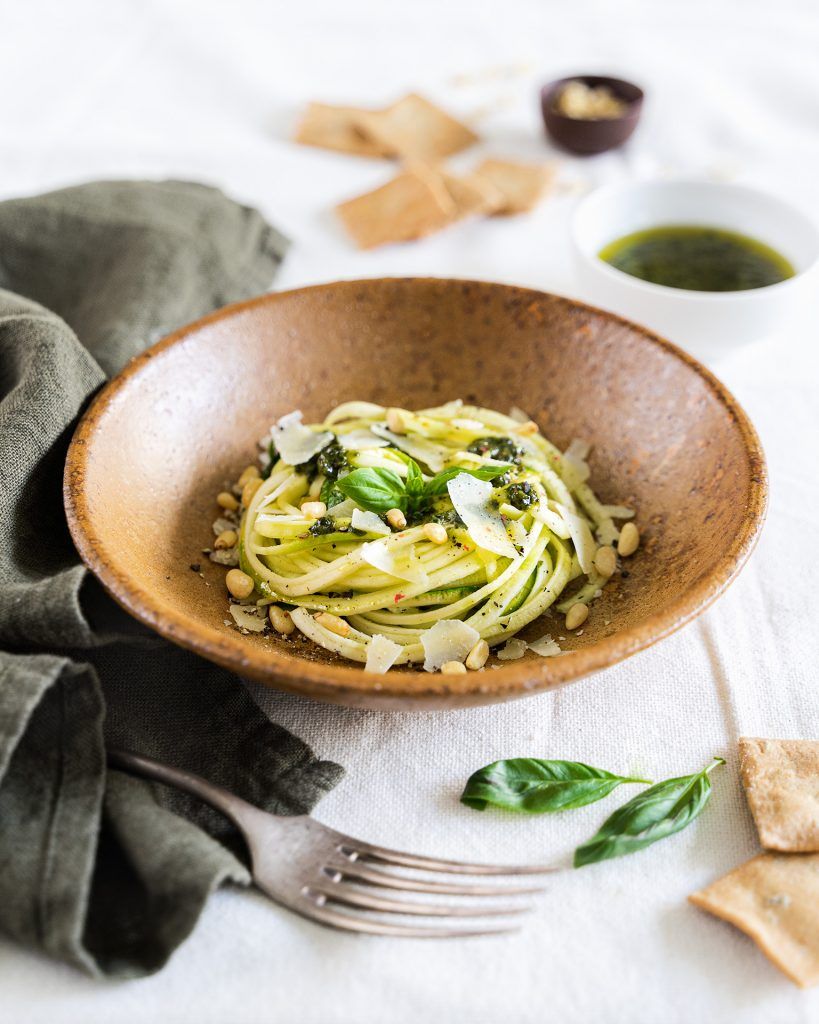Un bol de couleur terre cuite plein de spaghetti au pesto avec des courgettes crues, d'apparence délicieuse. Autour du plat, il y a des bols avec la sauce qui les accompagne et quelques toasts. Ce plat combine la richesse des pâtes avec la fraîcheur et la texture croquante des courgettes crues.