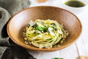 A clay-colored bowl full of pesto spaghetti with raw courgette, looking delicious. Around the dish, there are bowls with the accompanying sauce and some toast. This dish combines the richness of the pasta with the freshness and crunchy texture of the raw courgette.