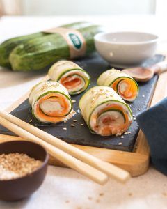 Quatre rouleaux de sushi faits avec de la courgette fraîche, du fromage à tartiner, du saumon, des graines et de la sauce soja, présentés sur une planche en ardoise avec des baguettes japonaises. Une recette rapide et délicieuse qui porte le sushi à un autre niveau.