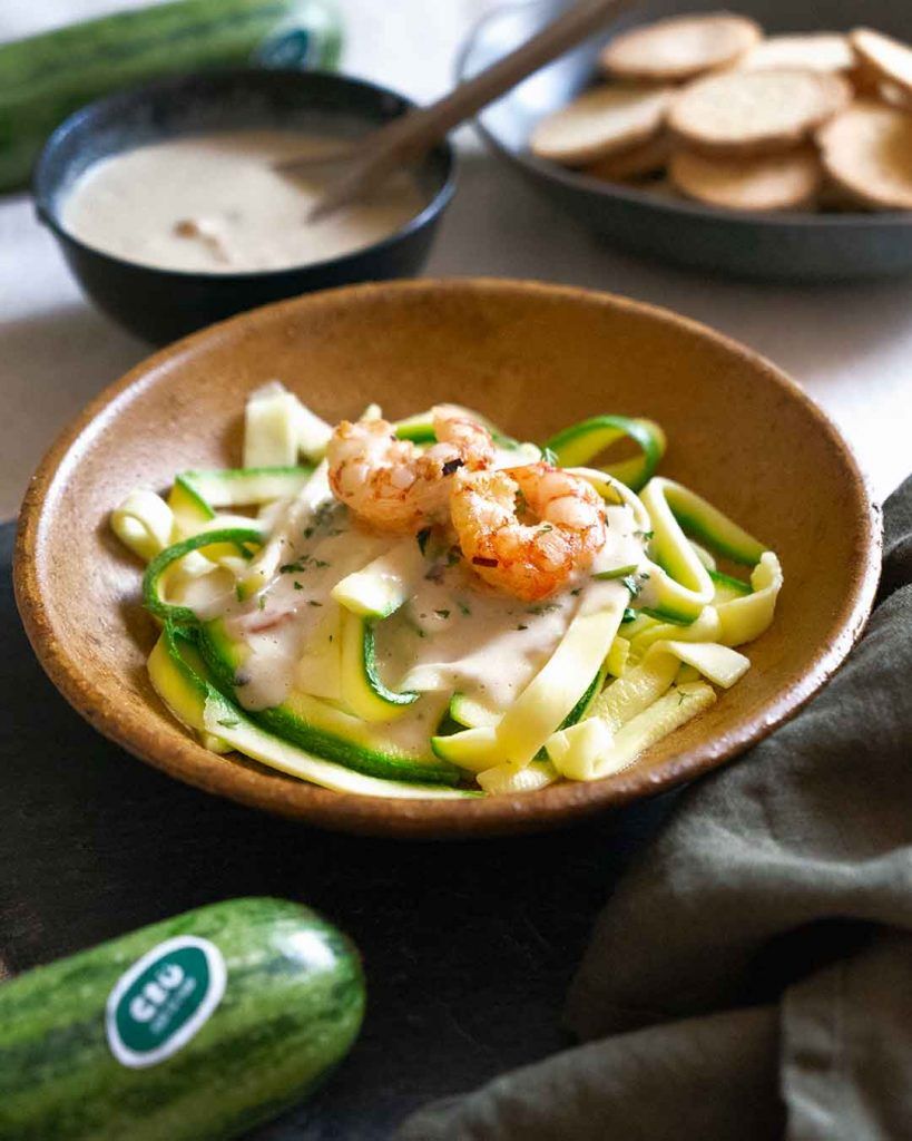 Un bol lleno de tallarines con gambas y calabacín crudo, con un aspecto delicioso. Alrededor del plato, hay cuencos con la salsa que los acompaña y algunas tostadas. Los tallarines son una forma crujiente y saludable de disfrutar de la pasta, sin renunciar al sabor.