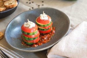 Dos pinchos con rodajas intercaladas de calabacín y tomate, coronados con burrata y aliñados con albahaca, presentados en un plato hondo de imitación piedra ovalado, sobre una salsa de tomate.