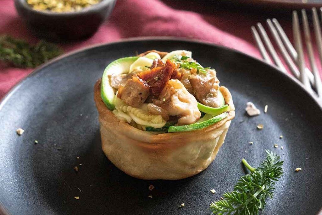 Une tartelette farcie avec la recette de Tulipes de champignons et courgette crue, présentée sur une assiette noire et décorée avec quelques herbes. Cette recette délicieuse et appétissante est parfaite pour surprendre votre famille, vos amis ou votre ex.